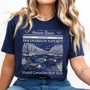 May include: Navy blue t-shirt with a graphic of a lake scene, trees, and loons. The text reads "Ontario Times" and "DISCOVERED IN NATURE!!!" with "Stupid Canadian Wolf Bird" below the image.