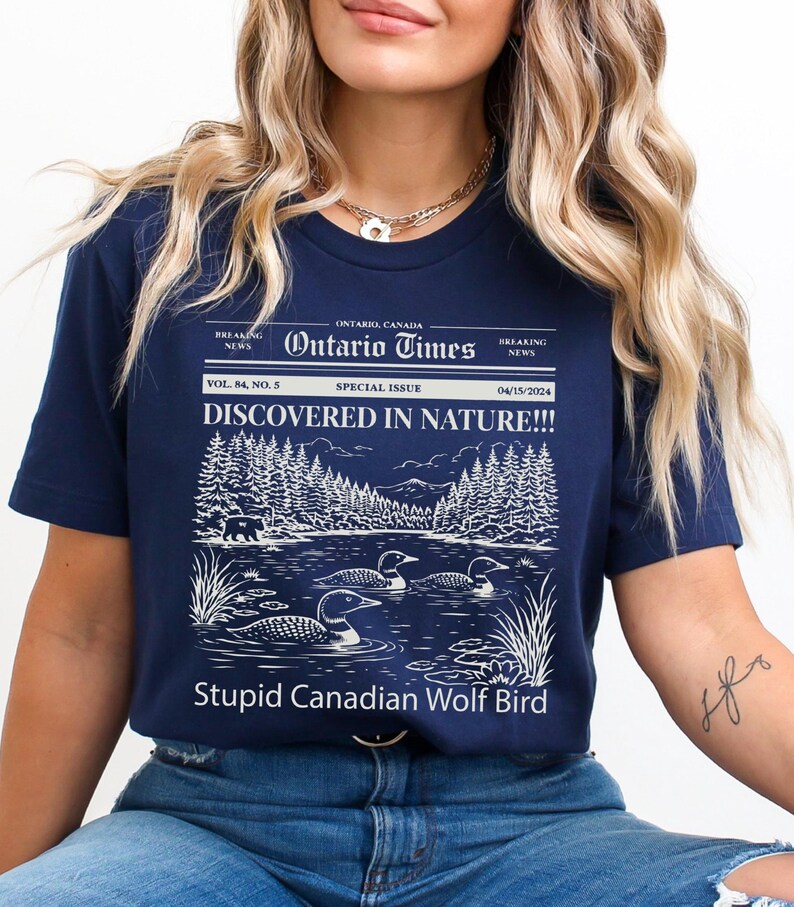 May include: Navy blue t-shirt with a graphic of a lake scene, trees, and loons. The text reads "Ontario Times" and "DISCOVERED IN NATURE!!!" with "Stupid Canadian Wolf Bird" below the image.