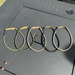 May include: Five slingshots with different coloured cords and wooden handles are displayed on a dark surface. The cords include camouflage, olive green, tan, black, and a tan and white pattern. These slingshots are designed for outdoor use.