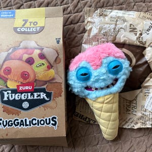 Könnte beinhalten: Ein Zuru Fuggler Fuggalicious Plüschtier in Form einer Eistüte mit rosa Oberteil, hellblauem Gesicht und gelber Waffelbasis. Das Plüschtier hat ein breites Grinsen mit sichtbaren Zähnen und schläfrigen Augen. Daneben steht ein brauner Karton mit Produkttext.