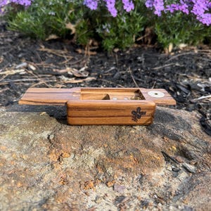 May include: A wooden box with a hinged lid, open to reveal a small compartment and a circular opening. The box is made of a light brown wood with a darker grain pattern. The box has a small logo on the front. Purple flowers are in the background.