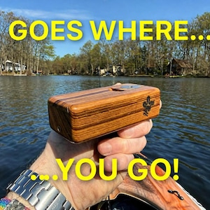 Puede incluir: Una caja de madera con un diseño de cactus se sostiene contra un fondo de lago. La caja tiene un patrón de vetas de madera a rayas. El texto "GOES WHERE... ...YOU GO!" está en letras amarillas en la parte superior e inferior de la imagen.