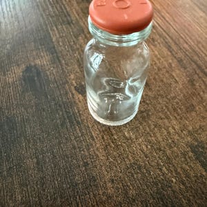 May include: A small, clear glass bottle with a reddish-orange cap. The bottle is cylindrical with a rounded base and a narrow neck. The cap has a circular design in the centre. The bottle is empty and sits on a dark brown wooden surface.