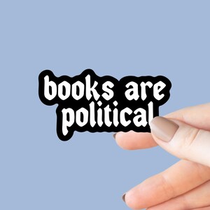 May include: A white sticker with the words "books are political" in a bold, stylized font with a black outline. The sticker is held by a hand against a light blue background.