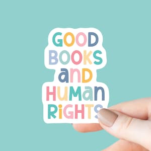 May include: A white sticker with the words "GOOD BOOKS and HUMAN RIGHTS" in colorful bubble letters. The sticker is held by a hand against a light blue background. The letters are in various colors, including blue, yellow, pink, and green.