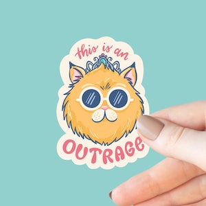 May include: A sticker featuring a cartoon cat with orange fur, wearing sunglasses and a crown. The sticker has the words "this is an OUTRAGE" in pink and white. The background is a light blue color.