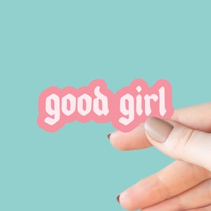 May include: A pink sticker with the words "good girl" in white, held by a hand against a teal background. The sticker has a light pink border and a rounded rectangular shape.