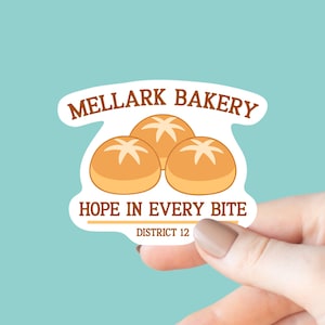 May include: A sticker featuring the text "MELLARK BAKERY" in brown, with three cartoon bread rolls. Below the rolls is the text "HOPE IN EVERY BITE" and "DISTRICT 12". The sticker is held against a teal background.