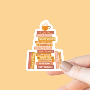 May include: A fall-themed sticker featuring a stack of books with phrases like "Hello Fall," "Sweater Weather," and "Happy Harvest." A cup of coffee with whipped cream sits atop the stack. The sticker is held by a hand against a light orange background.