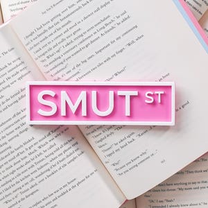 May include: A pink rectangular sign with the text "SMUT ST" in white, resting on open books. The sign's design is simple and modern, with a clean font.