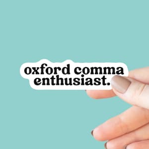 May include: A white sticker with the text "oxford comma enthusiast" in a bold, black font. The sticker is held by a hand with a light skin tone against a light blue background.