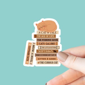 May include: A sticker featuring a stack of books with cat-themed titles, such as "A Cat's Tale" and "Soft Paws." A sleeping orange cat rests atop the books. The words "Meow: A Novel" and "Cottage Cats" are also visible.