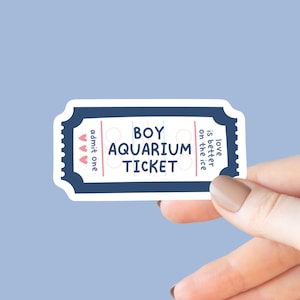 May include: A sticker in the shape of a ticket with the words "BOY AQUARIUM TICKET" in black. The ticket has a navy blue border with a scalloped edge. The words "admit one" and three pink hearts are on the left side.