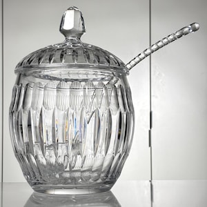 Può includere: Una zuccheriera trasparente in cristallo tagliato con coperchio e cucchiaio. La ciotola ha una forma arrotondata con tagli verticali strutturati. Il coperchio ha un pomello sfaccettato. Anche il cucchiaio è in cristallo tagliato.