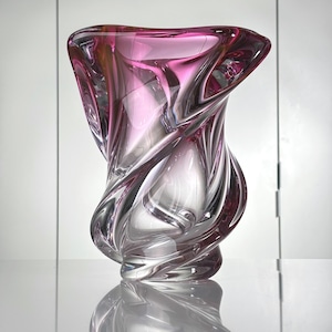 May include: A decorative glass vase with a swirling, organic design. The vase is primarily clear with a vibrant pink hue at the top, creating a gradient effect. The vase's form is reflected on the surface below.