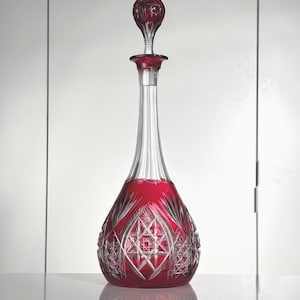 以下が含まれることがあります： ルビーレッドのクリスタルデカンタ。装飾的なストッパーが付いています。デカンタは長く透明な首と、模様の入ったファセットボディが特徴です。ストッパーは、お揃いのルビーレッドです。