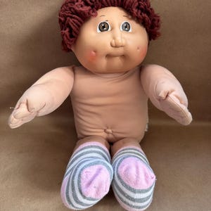 May include: A vintage doll with a soft, flesh-toned body, brown yarn hair, and painted facial features. The doll has rosy cheeks and is wearing striped socks in shades of pink, gray, and white. The doll's arms are outstretched.