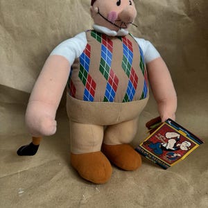 May include: A soft toy of a cartoon character wearing a burgundy beanie, a white collared shirt, and a brown argyle vest with red, blue, and green diamonds. The doll has tan legs and orange shoes, and holds a tag with cartoon characters.
