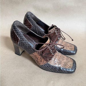 May include: A pair of vintage heeled shoes with a snakeskin pattern in shades of brown, tan, and grey. The shoes have a square toe, lace-up closure, and a chunky heel. The shoes are displayed on a neutral background.