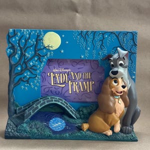 May include: A decorative photo frame featuring characters from Disney's "Lady and the Tramp." The frame has a blue background with a bridge, tree, and moon design. The title of the movie is displayed in the center.
