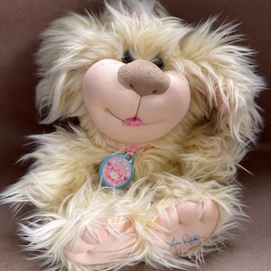 May include: A plush, cream-colored dog toy with a fluffy, shaggy appearance. It has a light brown nose, pink lips, and a blue tag that reads "CPK". The toy is signed in blue ink on one paw. A whimsical, soft toy.