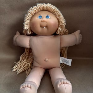 Peut inclure: Une poupée Cabbage Patch Kid vintage avec un corps en tissu doux et une tête en vinyle. La poupée a des cheveux blonds en fil, des yeux bleus et une petite bouche souriante. Elle n'est pas vêtue et porte une étiquette sur le côté.