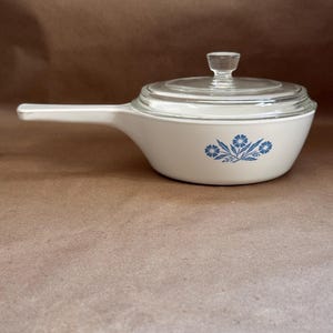 Peut inclure: Une casserole en céramique blanche avec un long manche et un couvercle en verre transparent. Le côté de la casserole présente un motif floral bleu composé de petites fleurs et de feuilles. Le couvercle possède un petit bouton en verre.
