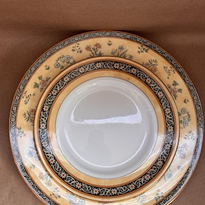 May include: A two-piece dinner set comprising a large and a smaller plate. The plates have a cream background with a floral pattern in orange, blue, and green. A black and gold border frames the inner white surface, creating an elegant look.