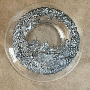 Peut inclure: Une assiette en verre transparent avec un motif de couronne argentée. La couronne présente des feuilles et des baies détaillées, entourant un oiseau détaillé. L'assiette est ronde et a un bord légèrement surélevé.
