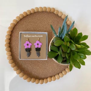 May include: A pair of handmade earrings with a floral design. The earrings feature a light purple flower, a black rectangular piece, and a gray leaf. The earrings are displayed in a white box with the text "Sofieczech.com". A succulent plant is next to the box.