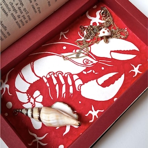 Peut inclure: Un plateau décoratif rouge et blanc avec un motif de homard, un coquillage et un collier pendentif champignon. Le plateau est dans une boîte rouge avec une page de livre visible. Le collier a une chaîne dorée.