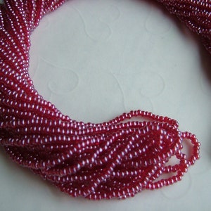 May include: A strand of small, red glass beads with a slight iridescent sheen. The beads are strung together on a white thread.