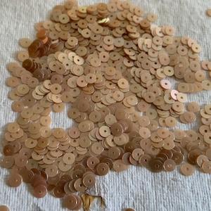 Puede incluir: Un montón de lentejuelas pequeñas, redondas, de color marrón claro, con un agujero en el centro. Las lentejuelas están dispersas sobre una superficie blanca y texturizada. Algunas lentejuelas tienen un ligero tono rosado.