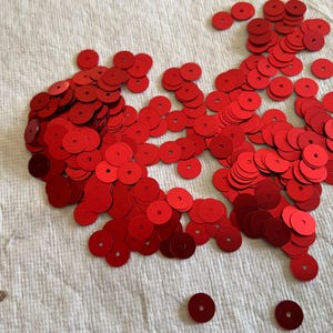 May include: A large pile of red sequins, scattered on a white fabric surface. The sequins are round and have a metallic sheen.