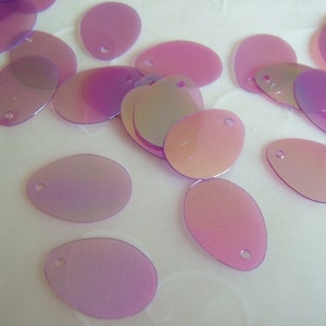 Puede incluir: Una dispersión de lentejuelas ovaladas iridiscentes de color morado claro. Las lentejuelas tienen un agujero en la parte superior para coser o sujetar.