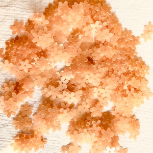 May include: A large pile of peach-colored flower-shaped sequins. The sequins are scattered on a white surface.