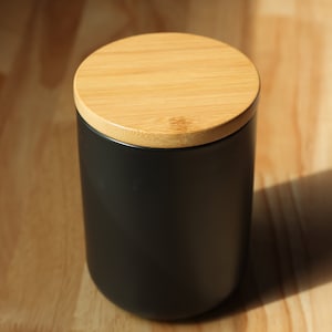 May include: A black ceramic container with a wooden lid. The container is cylindrical and has a smooth surface. The lid is made of natural bamboo and has a slightly textured surface.