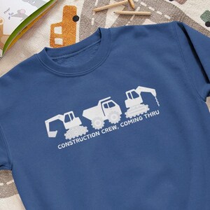 Peut inclure: Un sweat-shirt bleu marine avec un motif blanc de véhicules de chantier : une excavatrice, un camion benne et une chargeuse frontale. Le texte "CONSTRUCTION CREW. COMING THRU" est imprimé en dessous. Le sweat-shirt est sur un tapis à motifs.
