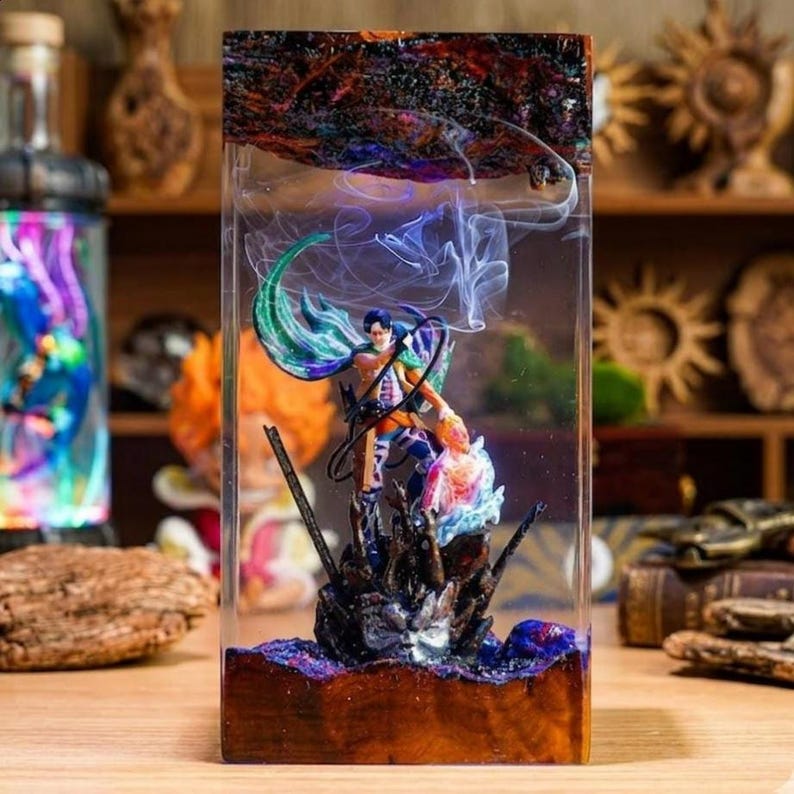 Puede incluir: Una pieza de arte de resina rectangular con una escena de figuras detallada. La obra de arte presenta una figura con una capa verde y azul, sobre un fondo de humo arremolinado e incrustada en un bloque de resina transparente. La base es de madera oscura.
