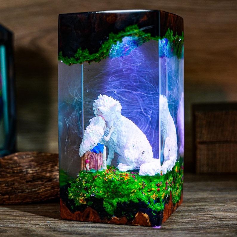 Puede incluir: Una pieza de arte de resina rectangular con una escultura blanca de un perro y una figura, dentro de un bloque de resina transparente. La base tiene un paisaje verde musgoso, y la parte superior tiene una veta de madera oscura. El interior tiene un efecto arremolinado azul y morado.