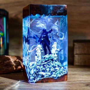 Pode incluir: Uma peça de arte em resina retangular com tema Star Wars. A obra apresenta Darth Vader e Stormtroopers em uma base azul texturizada, encapsulados em resina transparente com detalhes em espiral azul e branco. A parte superior e inferior são de madeira.
