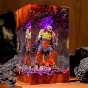 May include: A resin sculpture featuring a detailed action figure of a character in a yellow and orange armored suit. The figure is encased in a clear resin block with a wooden base and top, with purple and red accents.