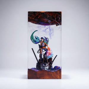 Puede incluir: Escultura rectangular de resina con dos figuras en una pose din&aacute;mica, encapsuladas en resina transparente. La escultura tiene una base de madera y una parte superior oscura y texturizada. Las figuras est&aacute;n detalladas con colores vibrantes, la resina tiene un efecto ahumado.