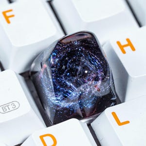 May include: A close-up of a black and blue galaxy-themed keycap on a white keyboard. The keycap is made of resin and has a swirling galaxy design with silver glitter. The keycap is on the 'H' key of the keyboard.