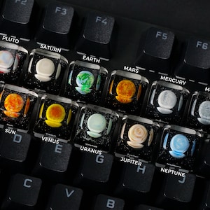 Pode incluir: Um teclado com teclas personalizadas que apresentam imagens dos planetas do nosso sistema solar. As teclas são transparentes e mostram os planetas de forma realista. As teclas são etiquetadas com os nomes dos planetas, incluindo Plutão, Saturno, Terra, Marte, Mercúrio, Lua, Sol, Vénus, Urano, Júpiter e Neptuno.