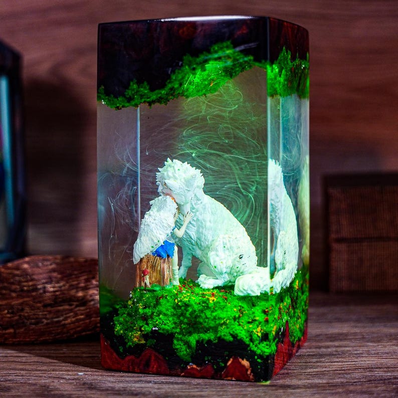 Puede incluir: Una escultura rectangular de resina con un perro blanco y una peque&ntilde;a figura abraz&aacute;ndose. La escultura tiene una base verde y roja con apariencia de bosque, y la parte superior es de color marr&oacute;n oscuro. El interior tiene un efecto et&eacute;reo y arremolinado.