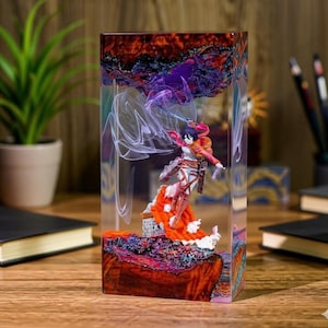 Puede incluir: Una pieza de arte rectangular de resina con una figura de un personaje en una pose din&aacute;mica. El bloque de resina tiene un dise&ntilde;o colorido y en capas con tonos rojos, naranjas y morados. La figura lleva un atuendo rojo y blanco.