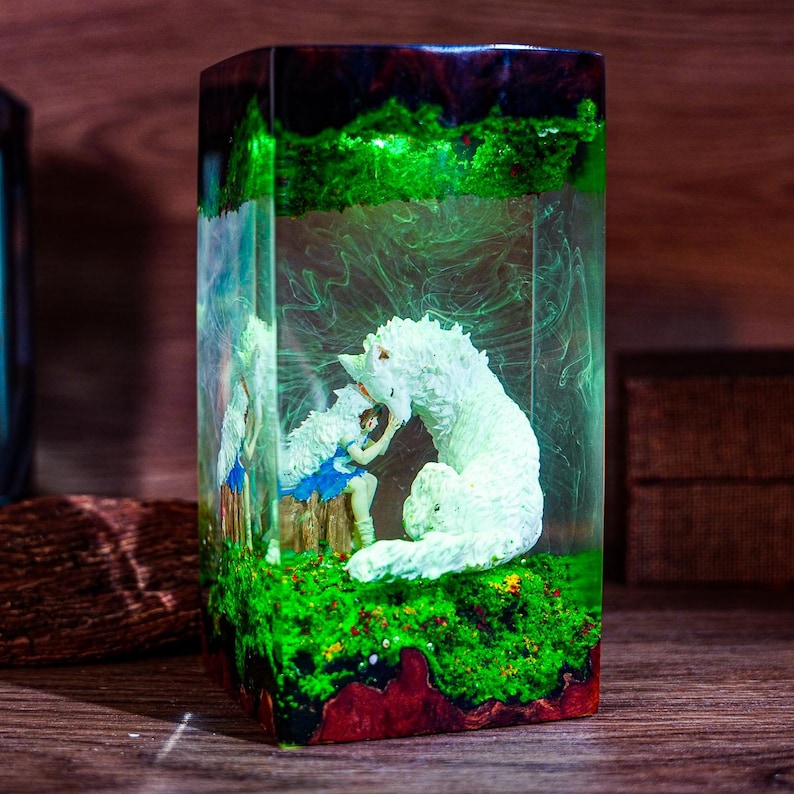 Puede incluir: Una escultura rectangular de resina con un lobo blanco y una ni&ntilde;a con un vestido azul, ambientada en un paisaje verde y rojo. La escultura est&aacute; encapsulada en resina transparente, creando una pieza de exhibici&oacute;n &uacute;nica.