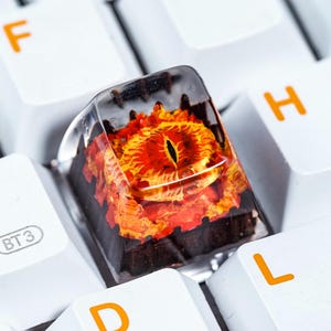 Könnte beinhalten: Nahaufnahme einer benutzerdefinierten Tastaturkappe mit einem roten und orangefarbenen Augenmotiv, das in klarem Harz gegossen ist. Die Kappe befindet sich auf einer weißen Tastatur.