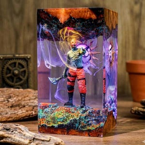 May include: A resin sculpture featuring a figure with an axe and a sun-like design. The figure is in red and orange pants and a blue shirt. The sculpture is encased in a clear resin block with wood grain edges and colorful accents.